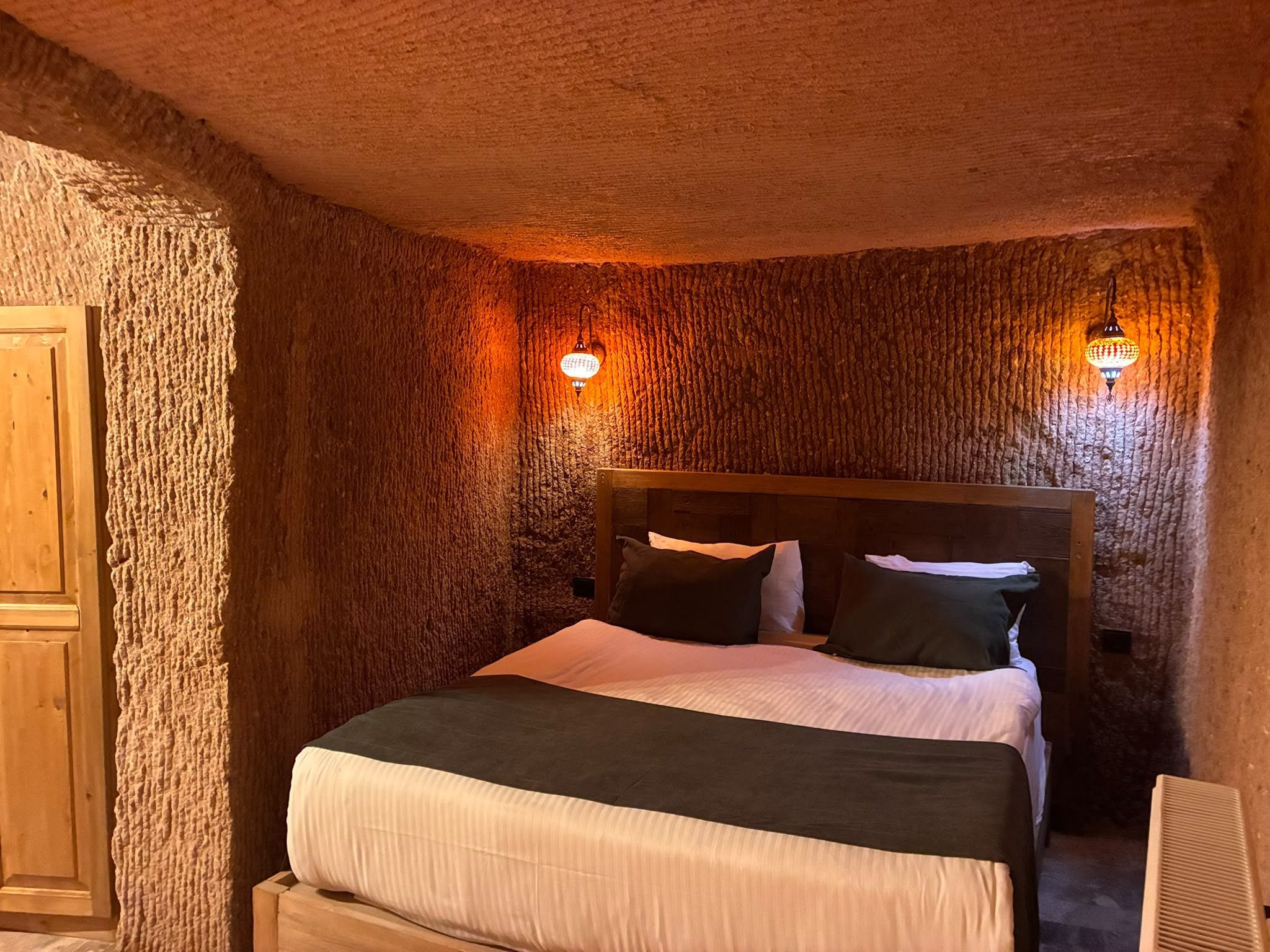Una habitación con pared de piedra texturizada, cama de madera con cabecera, dos lámparas fijadas a la pared y un armario de madera.