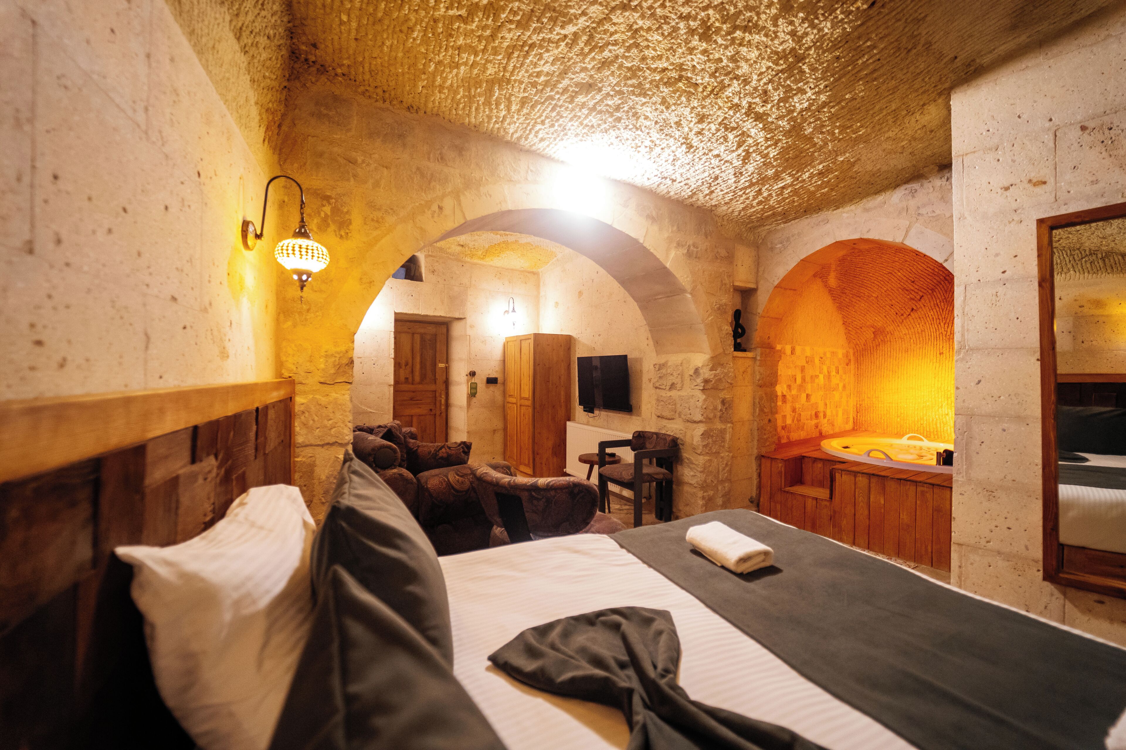 Una habitación con cama, cabecera de madera, una mesita, una silla, un banco de madera, televisión montada en la pared y pared de piedra.