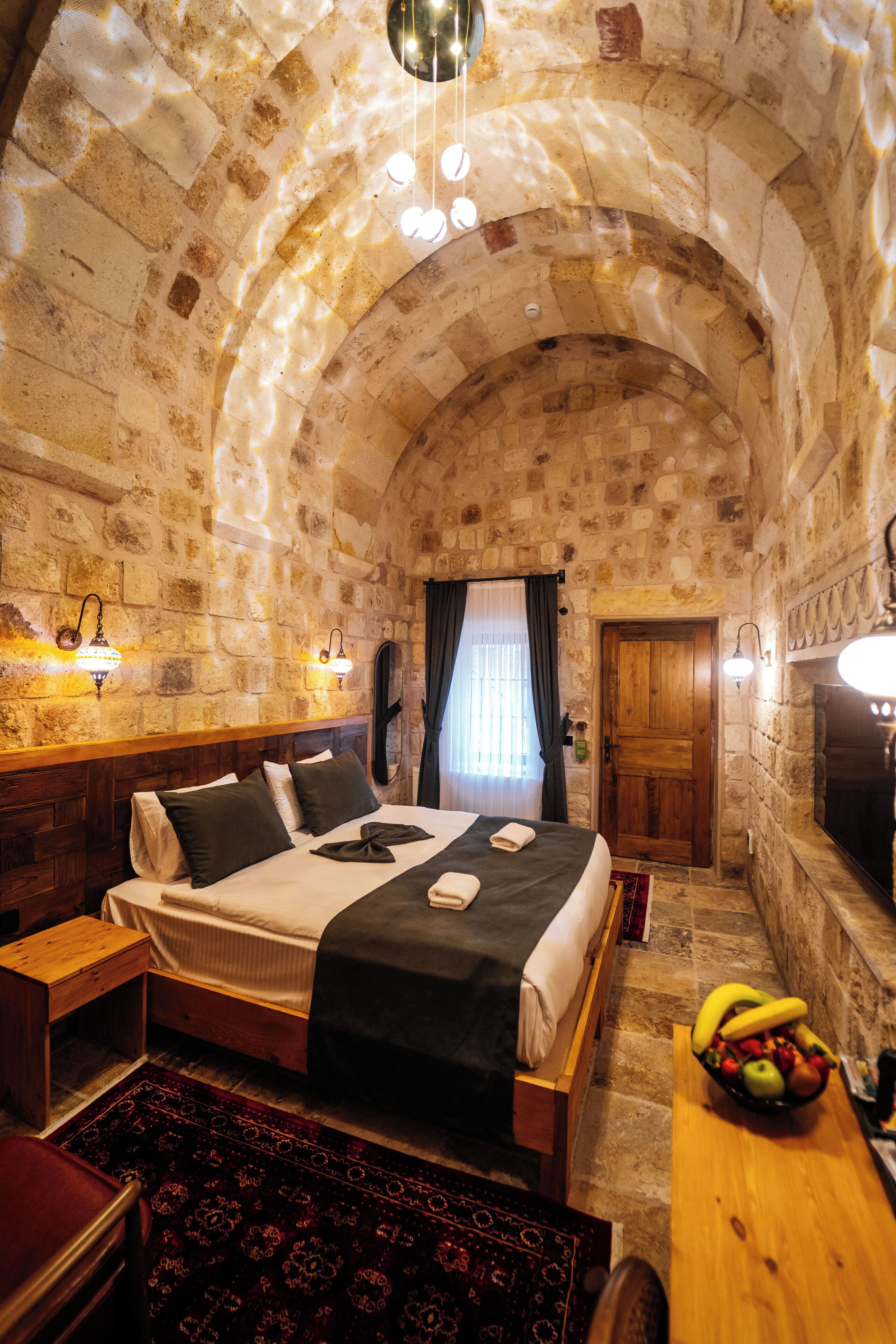 A stone-walled room with a bed, a wooden table with fruit, and a fireplace.