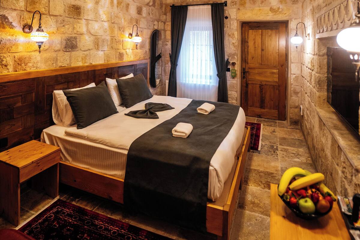 A stone-walled room with a bed, a wooden table with fruit, and a fireplace.