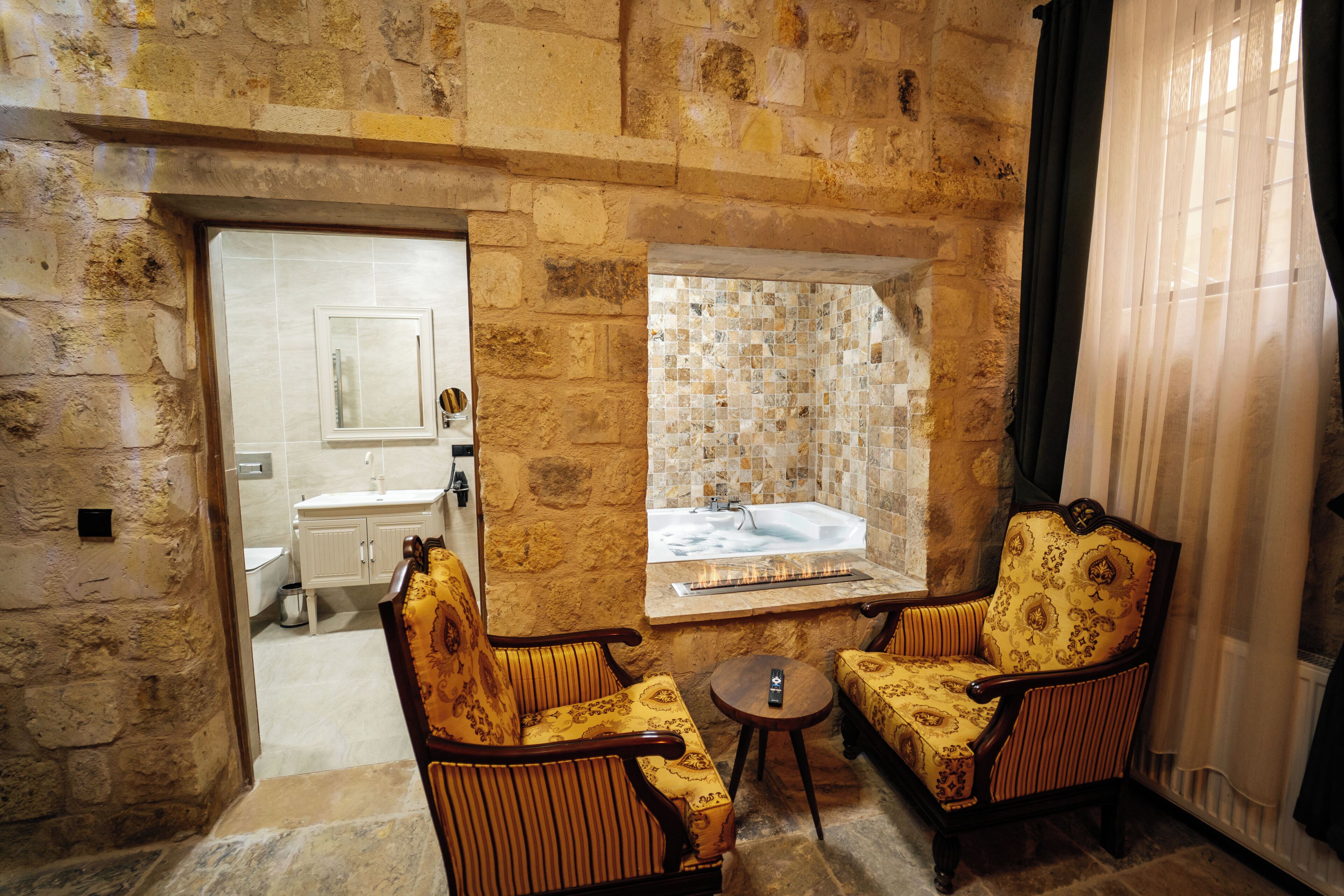 A stone-walled bathroom with a bathtub, two patterned armchairs, a small round table, and a window with a view of the bathroom.