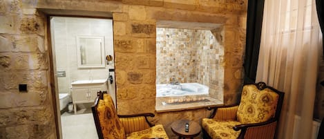 Ein Badezimmer mit Steinwänden, einer Badewanne, zwei gemusterten Sesseln, einem kleinen runden Tisch und einem Fenster mit Blick ins Badezimmer.