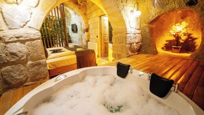 Ein Zimmer mit Steinwänden, einem hölzernen Bett, einer Badewanne voller Blasen und einem kleinen Tisch mit einem Stuhl.