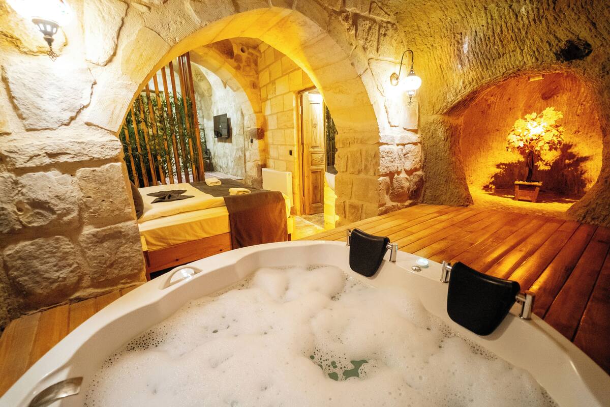 A stone-walled room with a wooden bed, a bathtub with bubbles, and a small table with a chair.