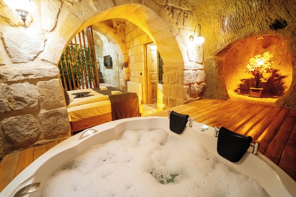 Quarto com paredes de pedra, uma cama de madeira, banheira com espuma e uma pequena mesa com uma cadeira.
