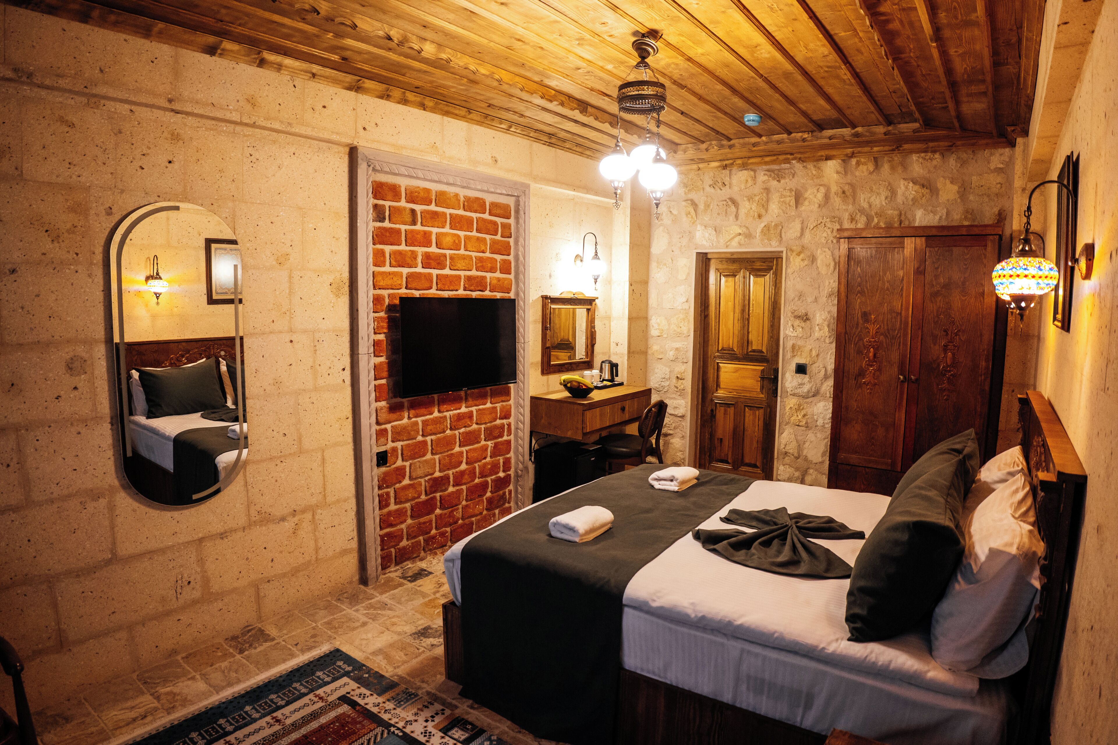 A bedroom with a brick fireplace, a bed with pillows, a desk with a chair, and a mirror.