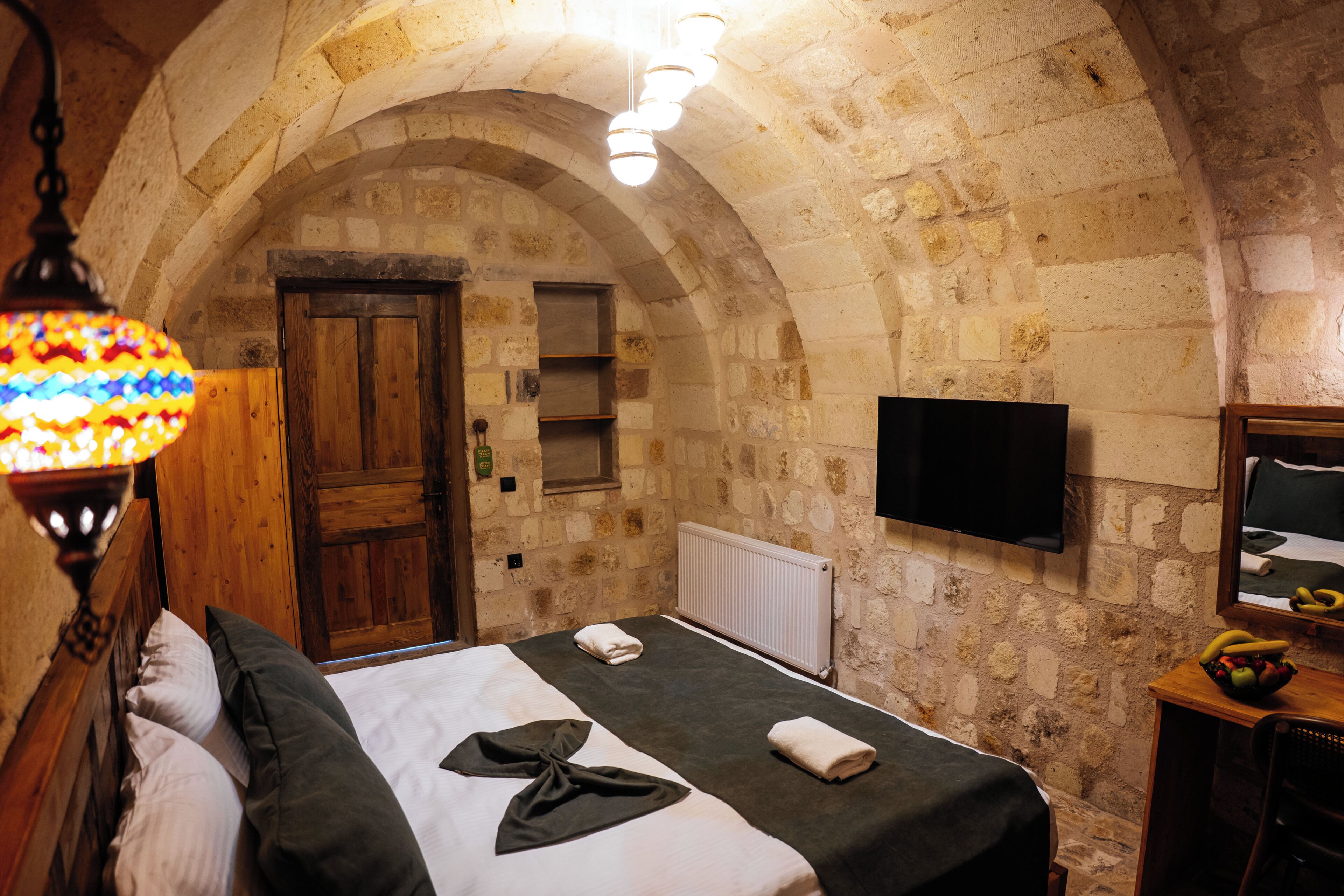 A room with a stone wall, a wooden door, a bed with a blanket, a wall-mounted TV, and a fruit bowl.