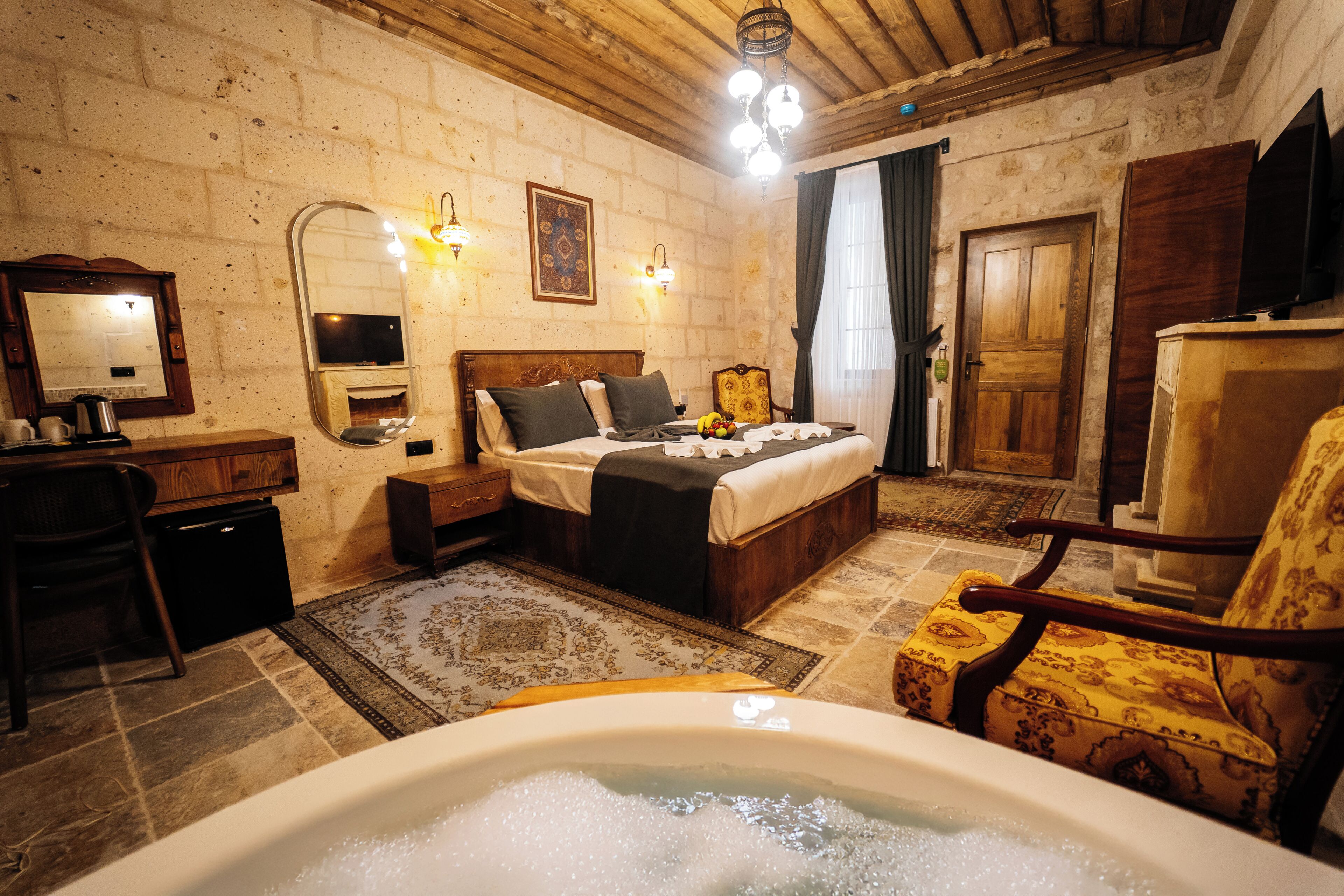 A room with a bed, a chair, a desk, a mirror, a television, and a bathtub.