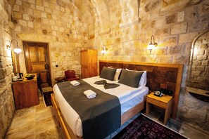 A stone-walled room with a bed, bedside table, lamp, and a wooden wardrobe.