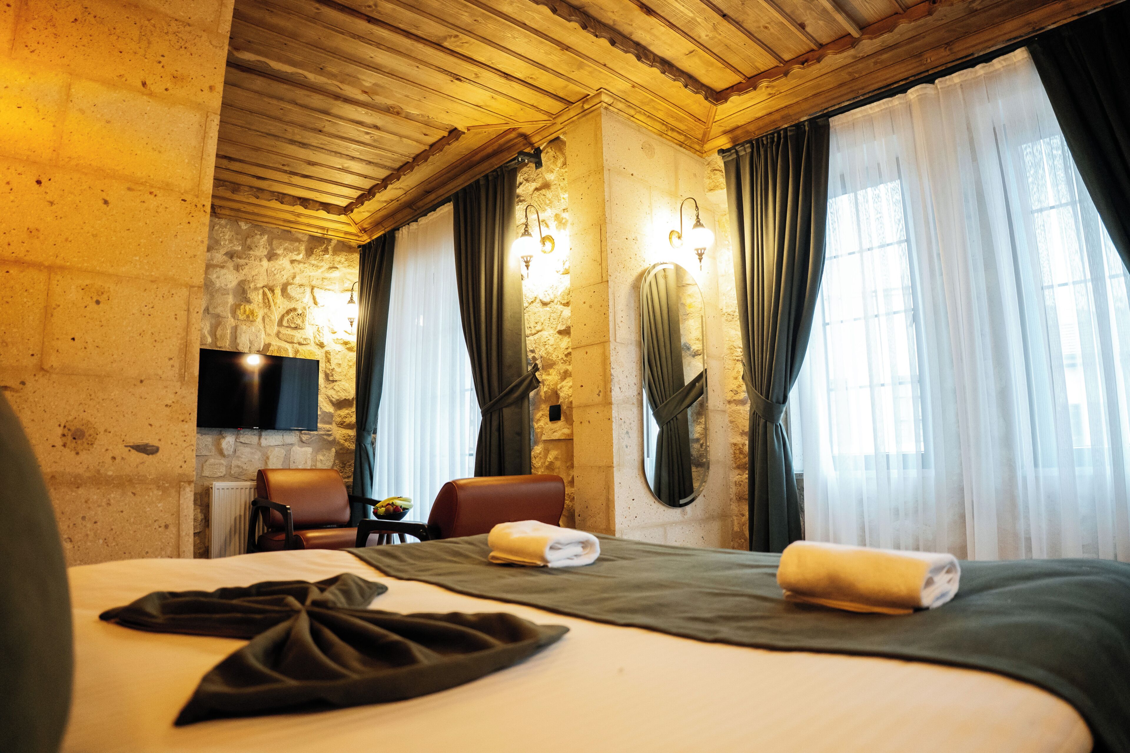 Habitación de hotel con pared de piedra, techo de madera, cama con colcha verde, sillón de cuero marrón, mesita con un bol de fruta y televisor de pantalla plana montado en la pared.