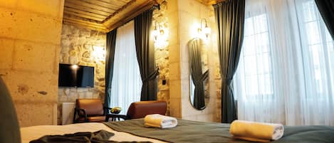 A hotel room with a stone wall, wooden ceiling, a bed with a green bedspread, a brown leather chair, a small table with a bowl of fruit, and a flat-screen TV mounted on the wall.