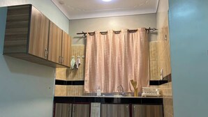 A compact kitchen with wooden cabinets, a sink, and a window with curtains. - Amala Homes – Private Stay in Mukono (Mukono)