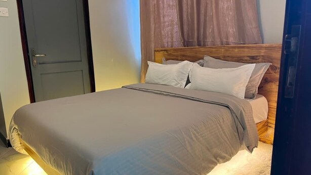 A bed with a wooden headboard, a grey bedspread, and a white pillow. - Amala Homes – Private Stay in Mukono (Mukono)