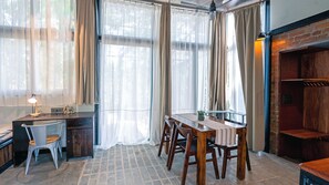 A room with a wooden dining table, chairs, a desk with a lamp, and large windows with curtains.