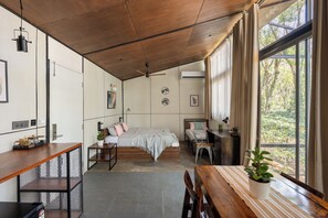 A modern room with a wooden ceiling, a bed with a grey blanket, a desk with a computer, and a large window with a view of trees.