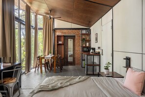 A modern room with a bed, desk, and dining area, featuring large windows and a brick wall.