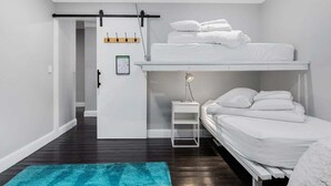 Um quarto com uma cama beliche, uma pequena mesa de cabeceira com luminária, e uma porta que dá para outro cômodo.