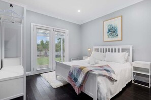 A bedroom with a white bed, a bedside table, a nightstand, a window with blinds, and a wall-mounted artwork.