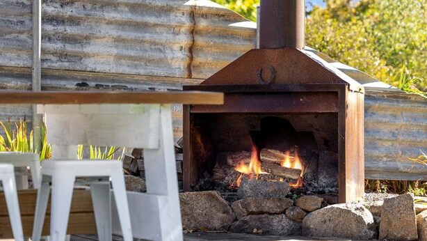Forno a lenha ao ar livre com chaminé de metal, rodeado por pedras e uma mesa de madeira.