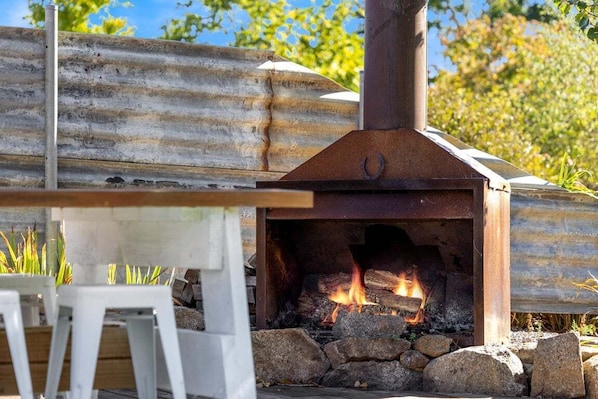 An outdoor wood-fired oven with a metal chimney, surrounded by rocks and a wooden table.