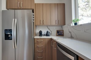 A kitchen with wooden cabinets, a stainless steel refrigerator, a marble countertop, a coffee maker, and a small potted plant. - Modern Home Near Old Mill & River (Bend)