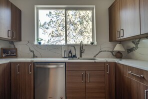 A kitchen with wooden cabinets, a stainless steel dishwasher, a marble countertop, a window with a view of trees, and a small potted plant. - Modern Home Near Old Mill & River (Bend)