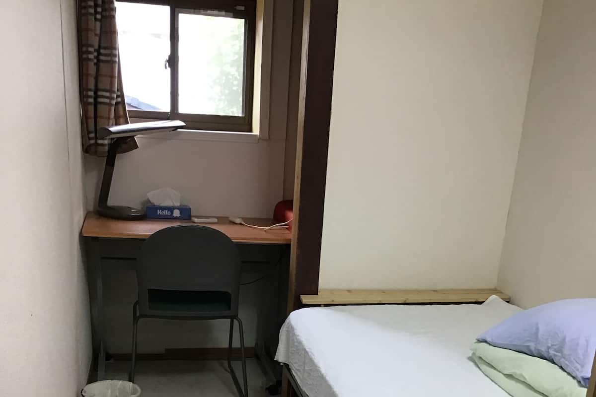A small room with a bed, a desk, a chair, a window, and a trash bin.