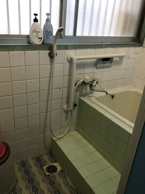 A bathroom with a bathtub, a shower, tiled walls, a window with blinds, and toiletries on the windowsill. - Chatamago (Nagasaki)