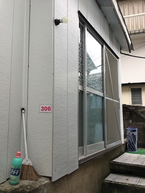 A building exterior with a number sign, a window with a patterned blind, a broom, and a cleaning bottle.