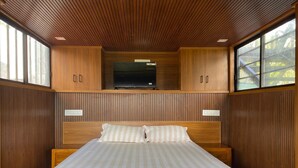 Un dormitorio con cabecera de madera, cama con ropa de cama a rayas, un televisor en un estante de madera y ventanas grandes.
