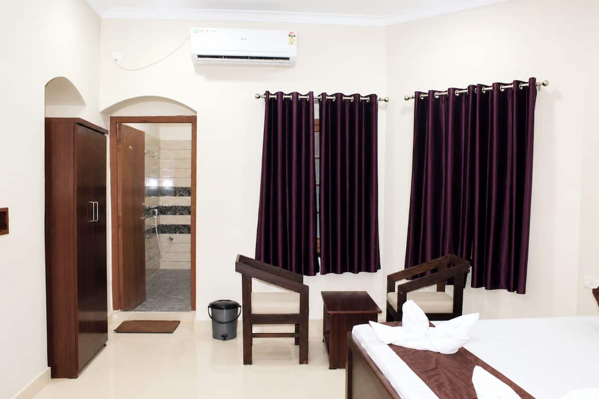 A hotel room with a bed, two chairs, a small table, a wooden wardrobe, a bathroom with a shower, and purple curtains.