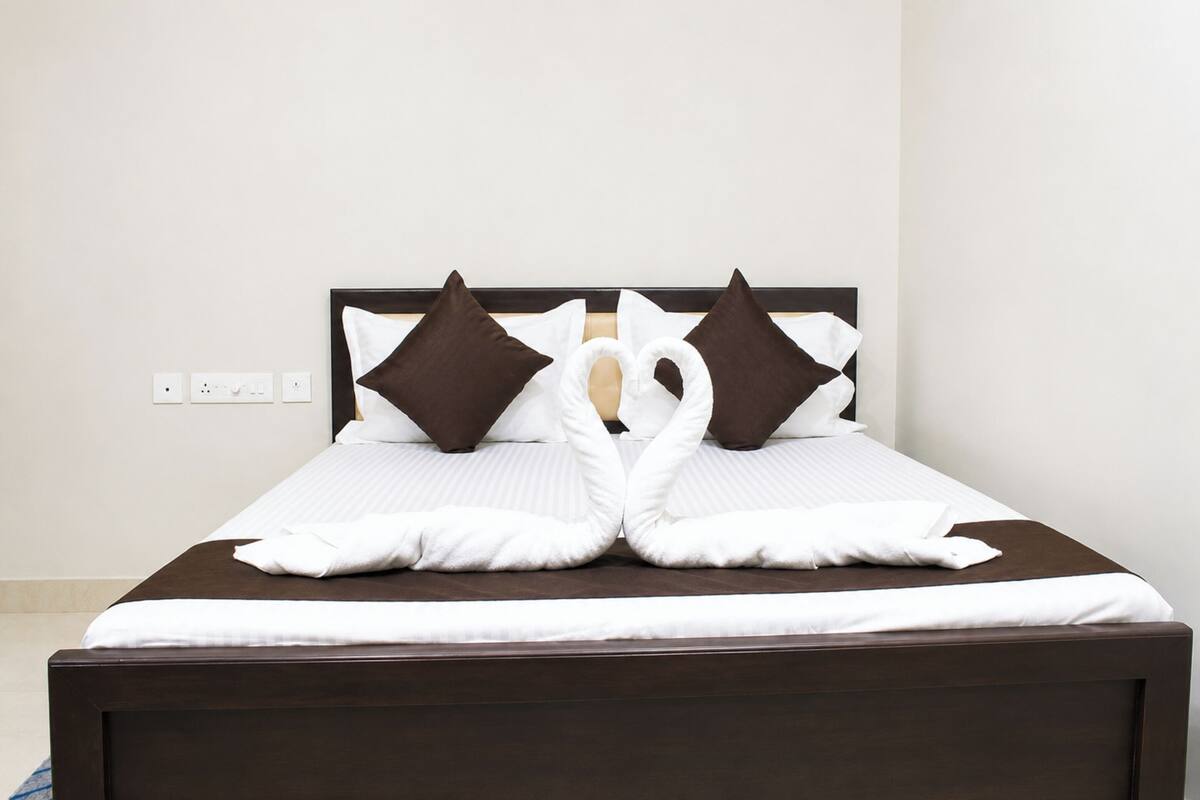 A neatly made bed with a heart-shaped pillow arrangement, a dark wooden headboard, and a plain wall with a power outlet.