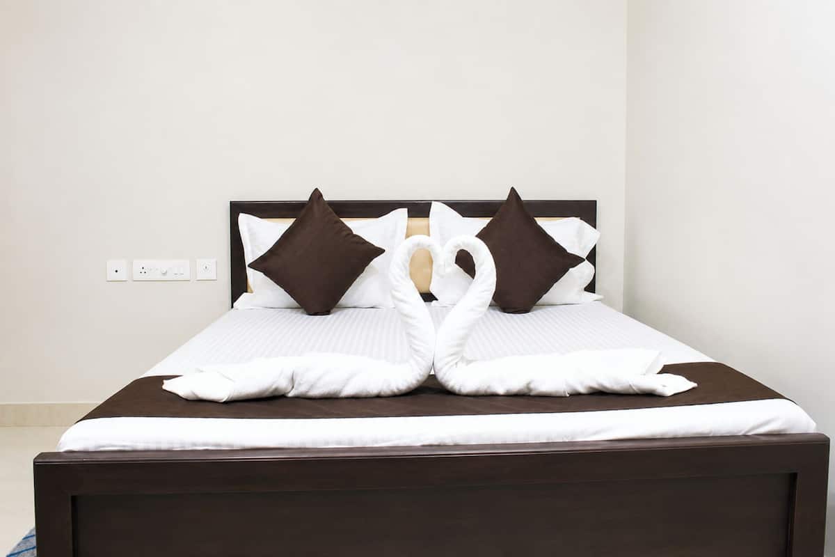 A neatly made bed with a heart-shaped pillow arrangement, a dark wooden headboard, and a plain wall with a power outlet.