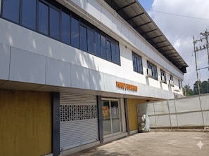 A two-story building with a closed shutter on the ground floor, a sign reading 'PIJOS & MESTERIA,' and a covered walkway on the upper level. - Hotel O Erumeli Bus Terminal Kottayam (Kanjirappally)