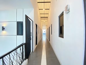 A long, narrow hallway with doors on both sides, a metal railing on the left, and a ceiling with recessed lighting. - Tamastu House Lippo Karawaci (Curug)