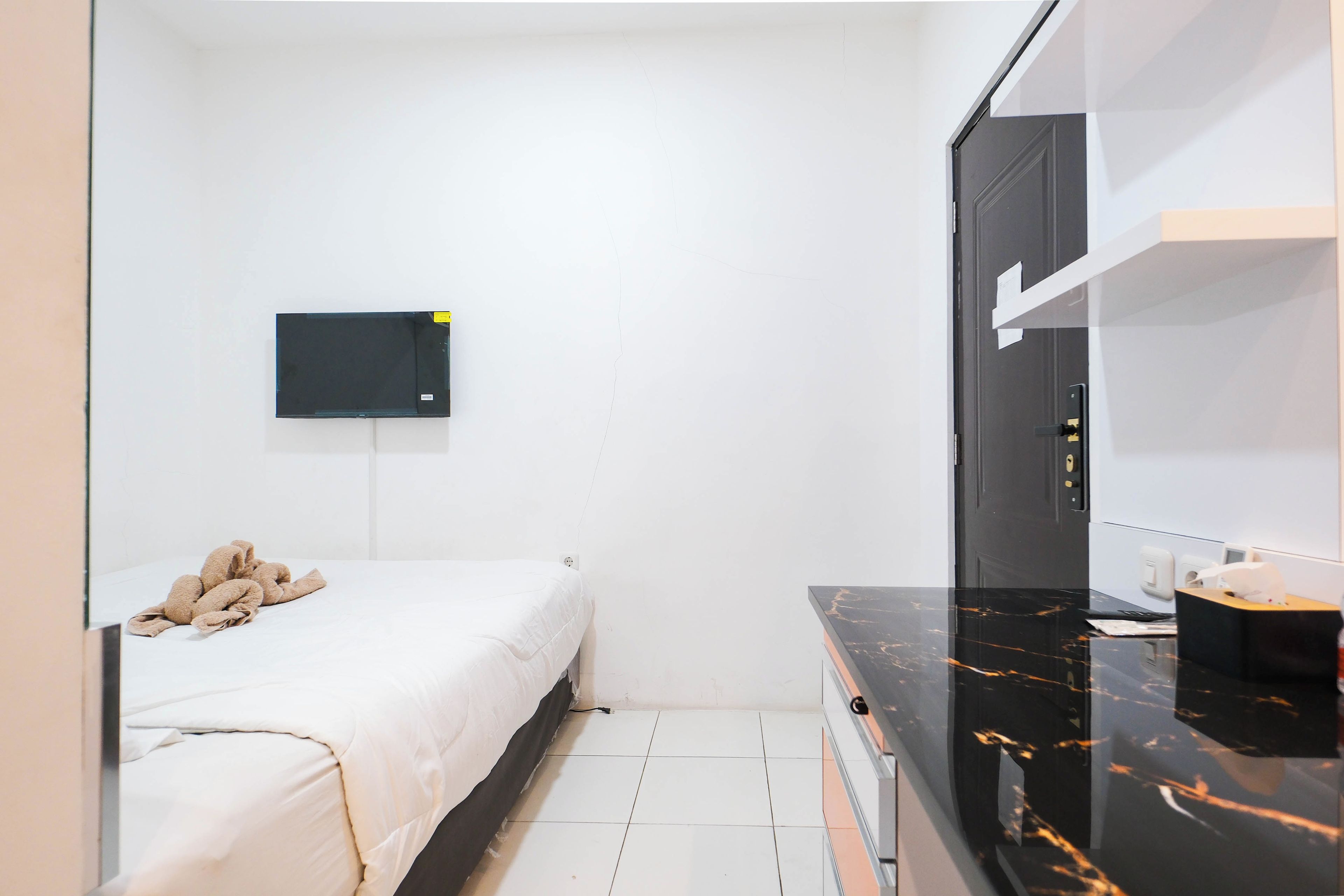 A small, modern bedroom with a bed, a flat-screen TV mounted on the wall, a mirror, and a small desk with a telephone.