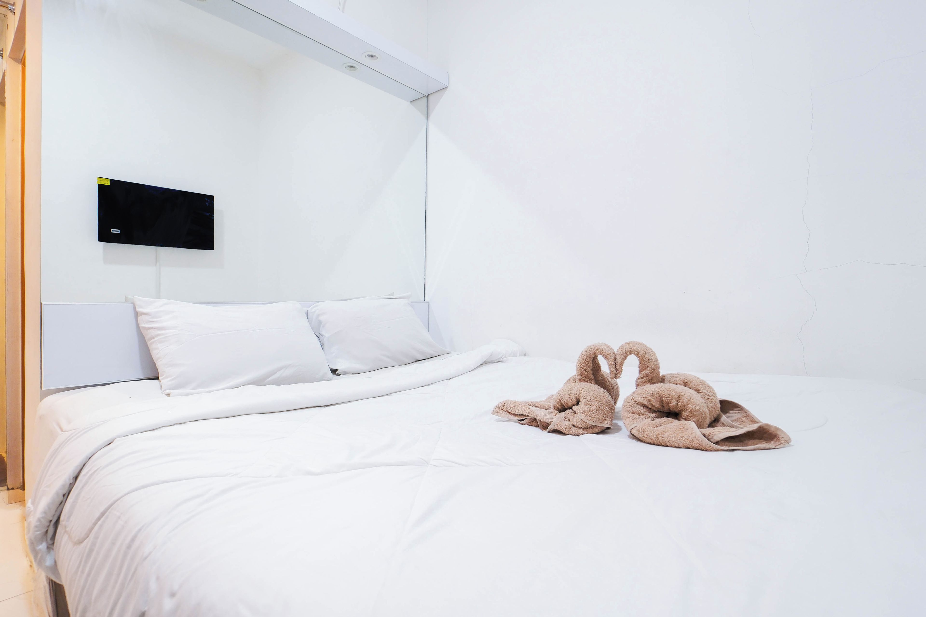A hotel room with a bed, white bedding, a flat-screen TV mounted on the wall, and a pair of brown slippers on the bed.