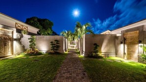 A nighttime view of a courtyard with a brick pathway, palm trees, and a building with a wooden door. - Bingin Villas: Luxury, Concierge &  Security (Badung)