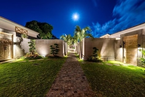 A nighttime view of a courtyard with a brick pathway, palm trees, and a building with a wooden door. - Bingin Villas: Luxury, Concierge &  Security (Badung)
