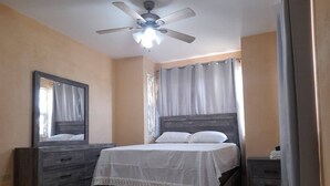 A bedroom with a bed, a dresser, a nightstand, a ceiling fan, and a window with curtains. - TJ Guest House - Jamaica (Spanish Town)