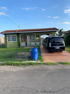 A house with a tiled roof, a parked car, and a blue barrel. - TJ Guest House - Jamaica (Spanish Town)