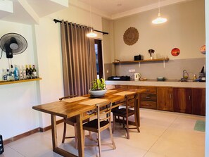 Un comedor con mesa y sillas de madera, un ventilador, una planta en maceta y una cocina al fondo.