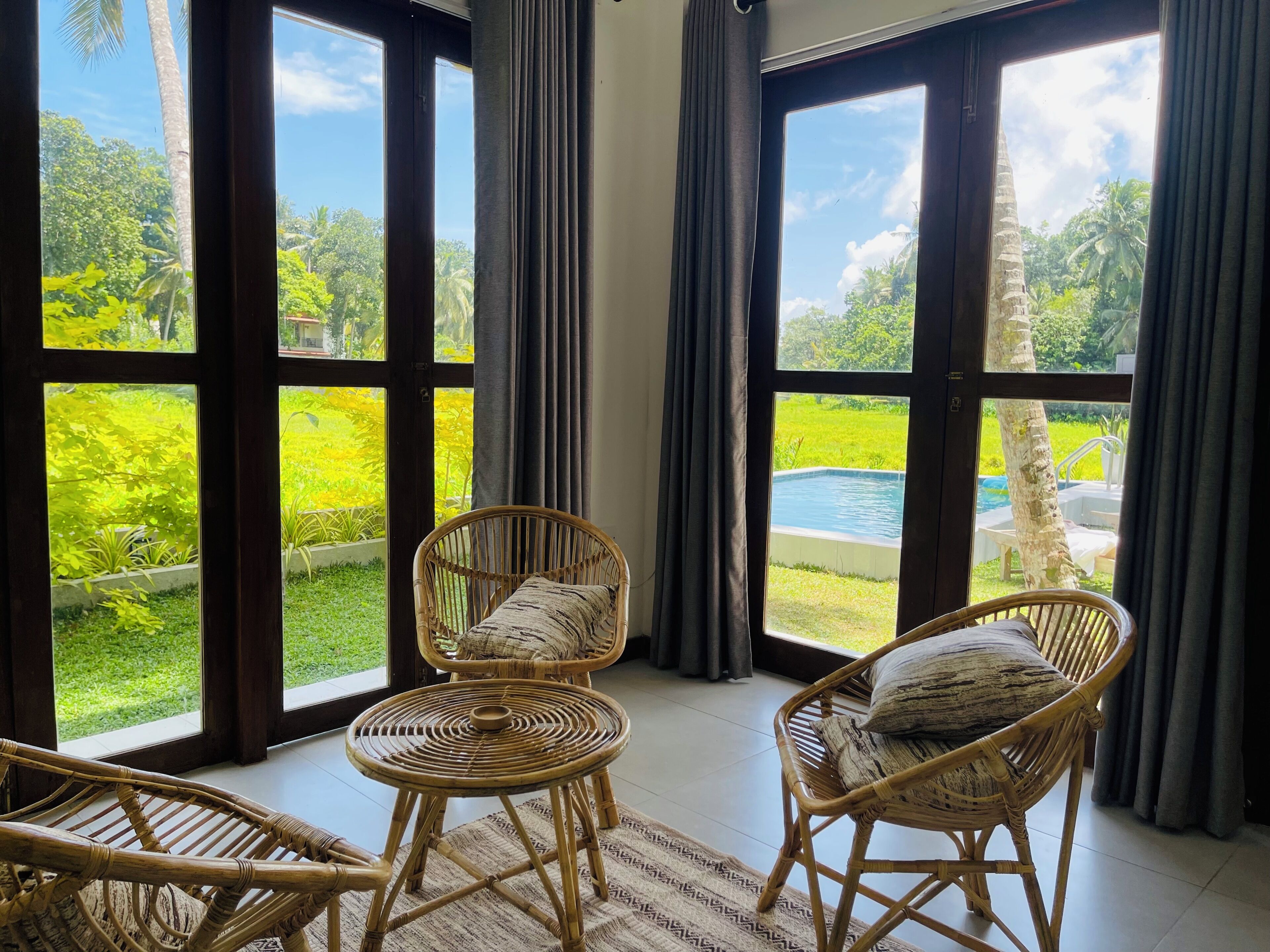 A room with large windows, wicker furniture, and a view of a swimming pool and greenery.