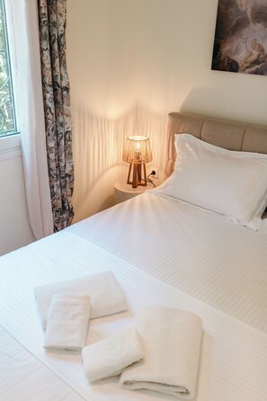 Una cama bien hecha con sábanas y almohadas blancas, una lámpara de noche y una ventana con cortinas.