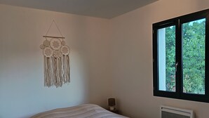 Ett sovrum med en säng, en väggdekoration i makramé, ett sängbord med en lampa och ett fönster med utsikt över grönområden.