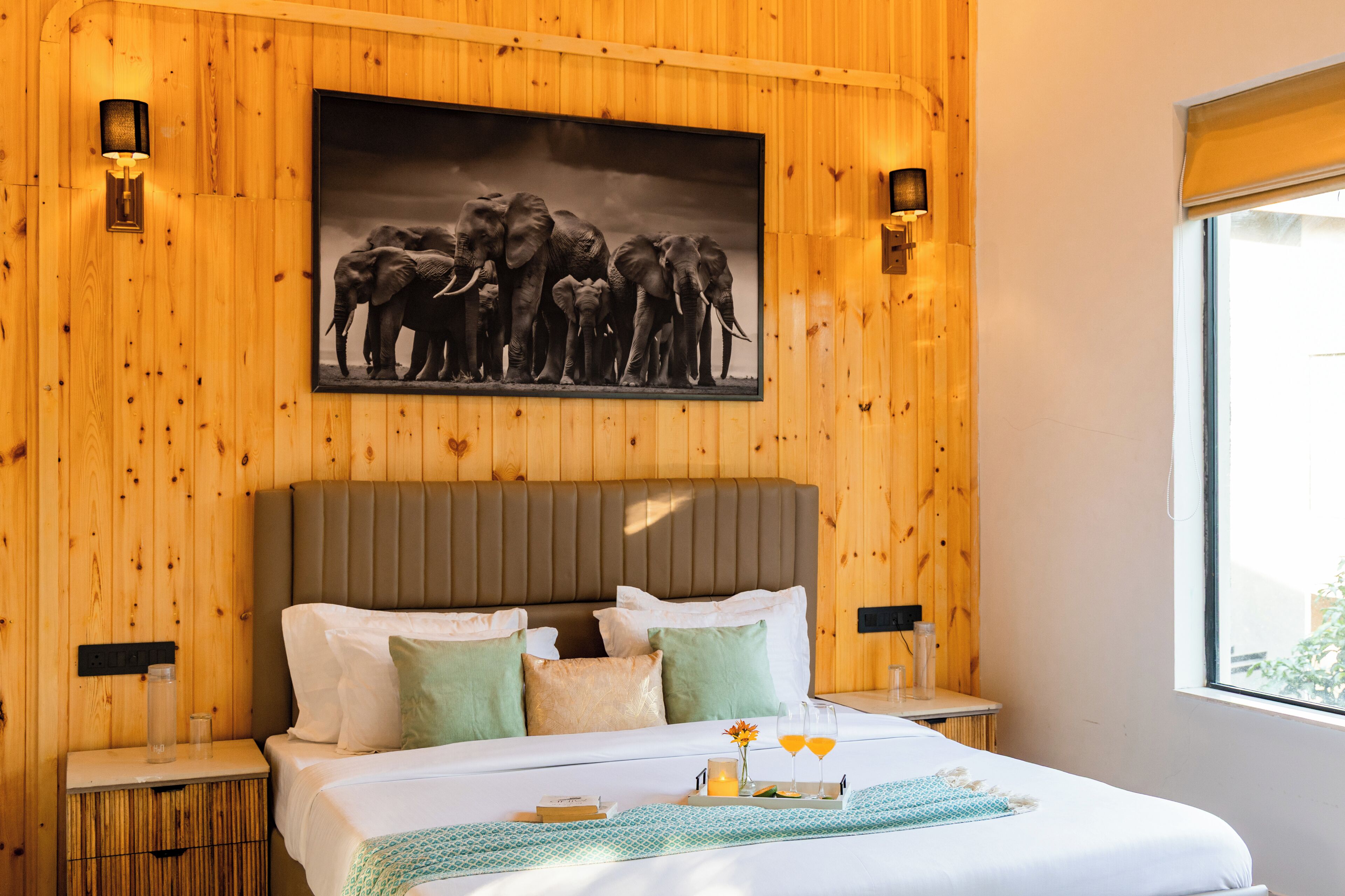 A wooden-paneled room with a bed, pillows, a tray with glasses and a bottle, and a framed picture of elephants.