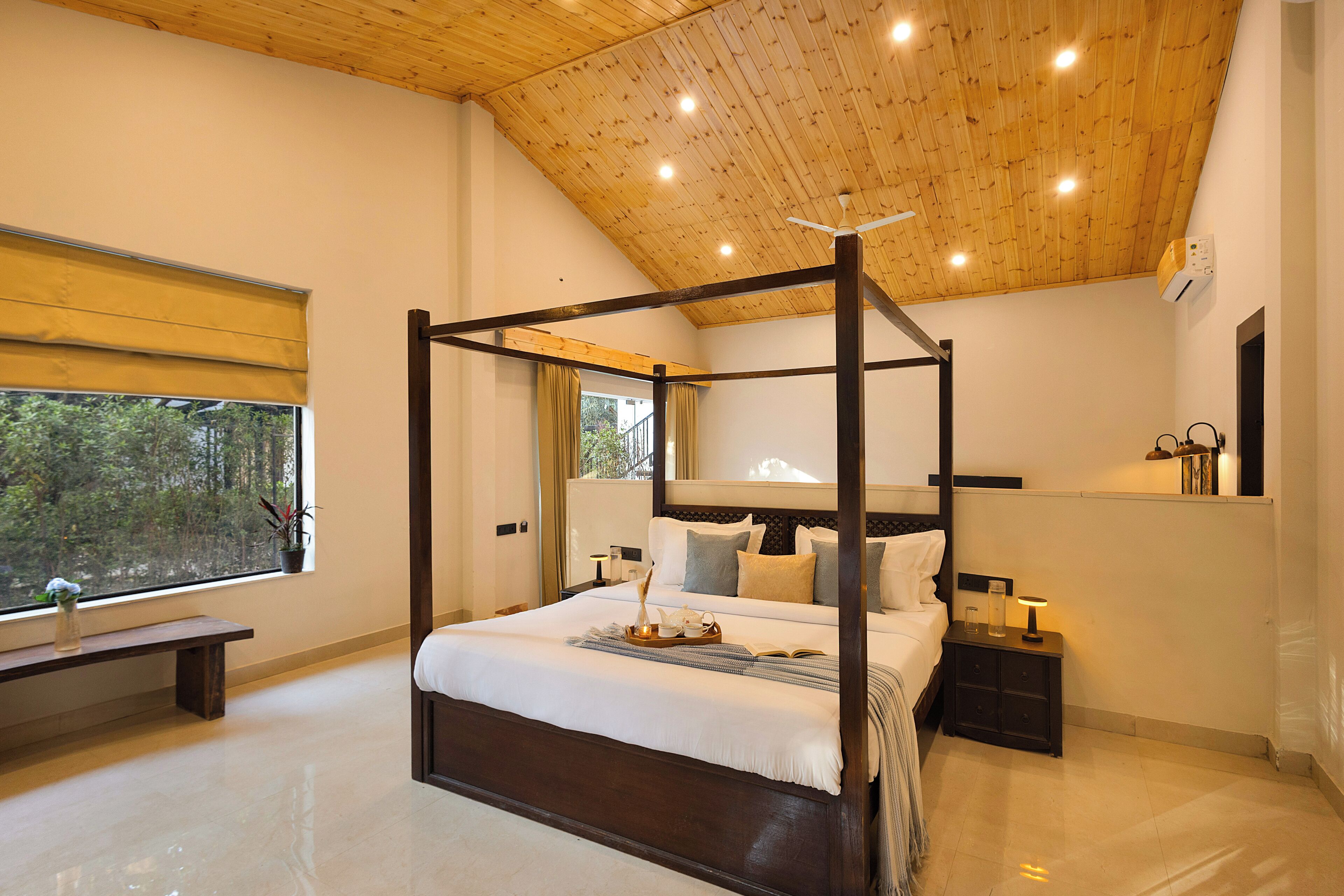 A four-poster bed with a wooden headboard, a bedside table with a lamp, a bench by the window, and a view of trees outside.