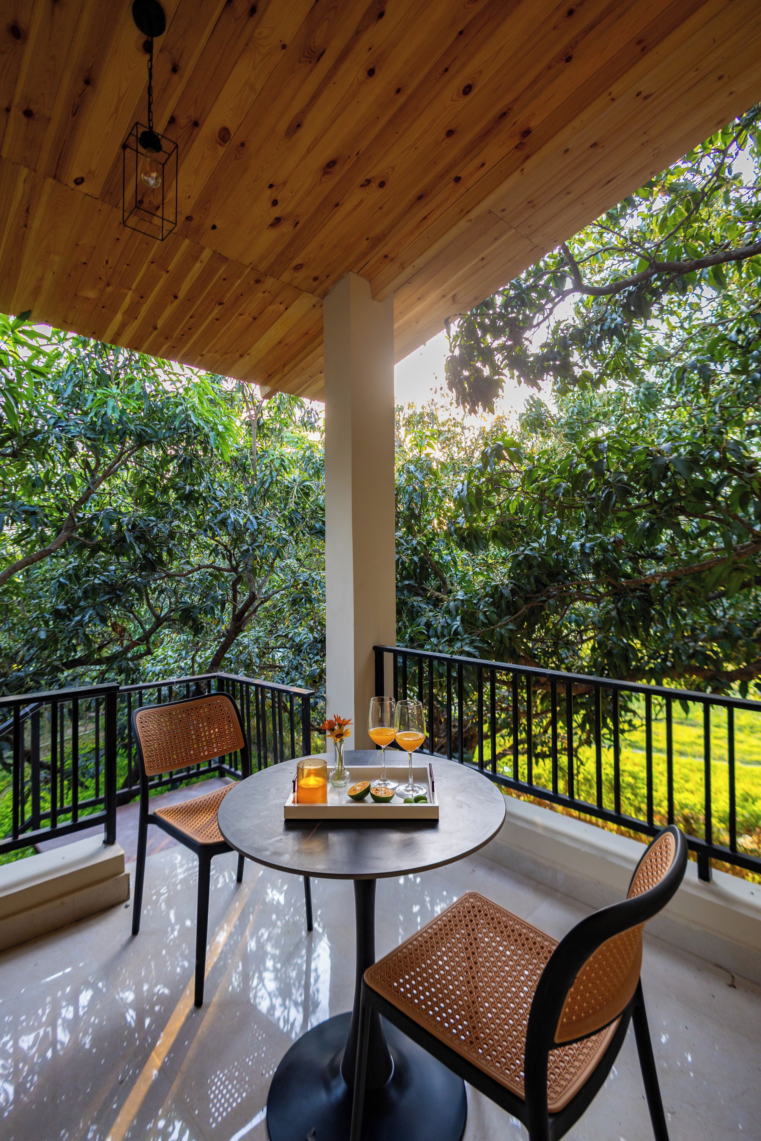A wooden deck with a round table and two wicker chairs, surrounded by lush greenery.