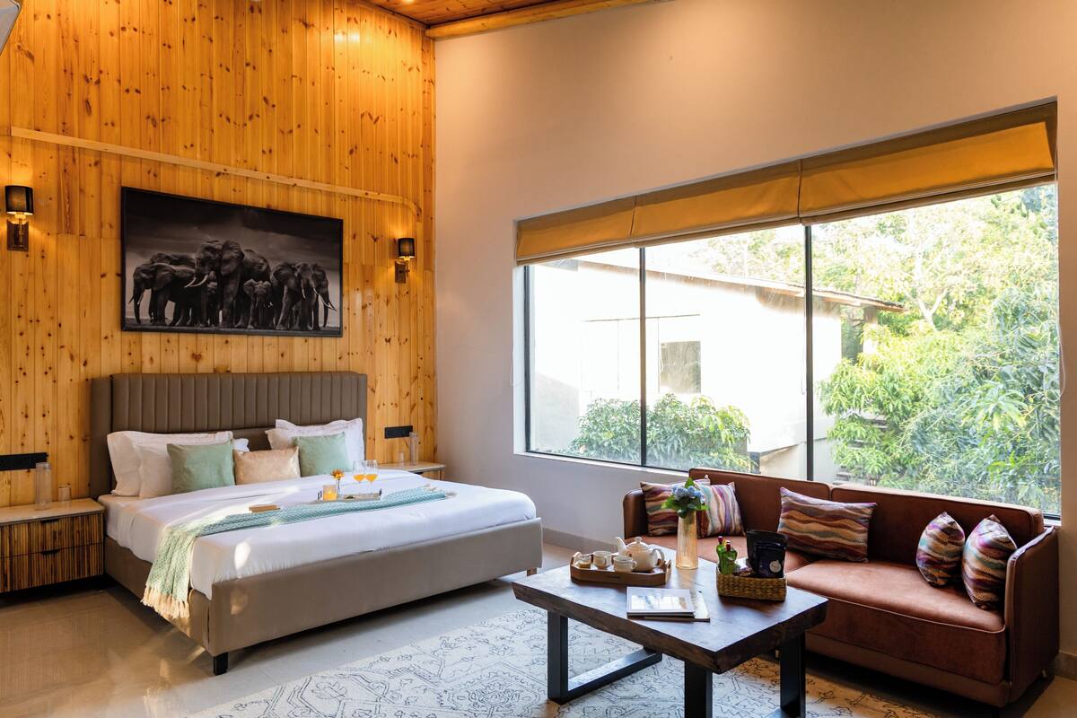 A bedroom with a bed, a sofa, a coffee table, a painting, and a window with a view of greenery.