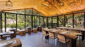 A restaurant with wooden tables and chairs, large windows, and a view of greenery. - Sylvaara Suites by ELIVAAS Jim Corbett (Ramnagar)